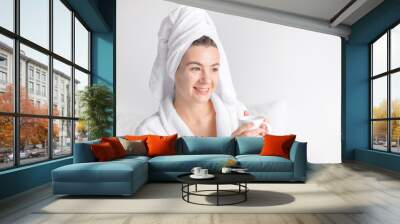 a beautiful young woman in a bathrobe and with  bath towel on her head sits on the bed and holds a white cup of coffee at home in front of the window  Wall mural