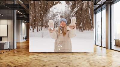 Happy fashionable young woman tossing snow up in frosty winter park wearing stylish warm clothes and having fun outdoors. Seasonal activities. Space Wall mural