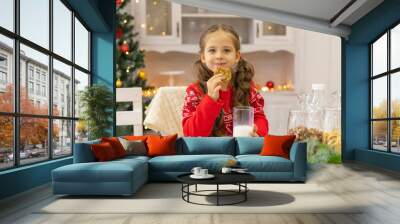 Happy joyful cute little girl in red sweater drinking milk and eating delicious cookie near Christmas tree in the kitchen for Christmas morning. Christmas time. Christmas holidays Wall mural