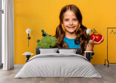 Happy little girl standing with green raw broccoli and tomatoes over yellow background. Healthy food for children. Little girl with eco vegetables, isolated, space for text, concept organic food Wall mural