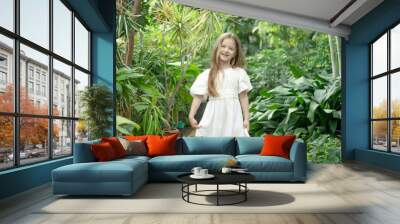 Little blonde girl in the botanical garden. a girl in a white dress exploring tropical plants and flowers in the greenhouse. Wall mural