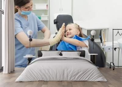 Little patient girl sitting in a chair gives a high five to a pediatric dentist after dental treatment at a clinic with modern equipment. Concept of modern and painless treatment of children's teeth. Wall mural