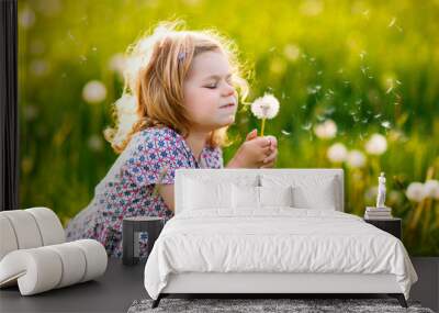 Adorable cute little baby girl blowing on a dandelion flower on the nature in the summer. Happy healthy beautiful toddler child with blowball, having fun. Bright sunset light, active kid. Wall mural