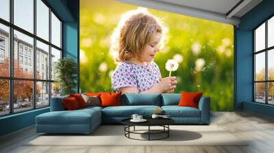 Adorable cute little baby girl blowing on a dandelion flower on the nature in the summer. Happy healthy beautiful toddler child with blowball, having fun. Bright sunset light, active kid. Wall mural