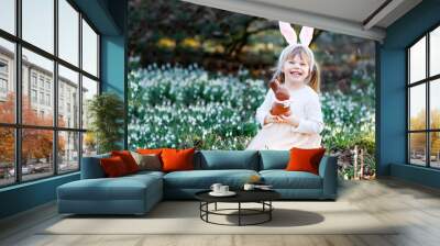 adorable little girl with easter bunny ears eating chocholate figure in spring forest on sunny day,  Wall mural