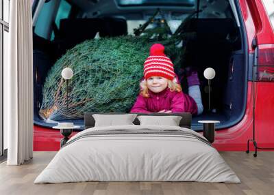 Adorable little toddler girl with Christmas tree inside of family car. Happy healthy baby child in winter fashion clothes choosing and buying big Xmas tree for traditional celebration. Wall mural