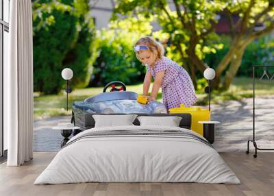 Cute gorgeous toddler girl washing big old toy car in summer garden, outdoors. Happy healthy little child cleaning car with soap and water, having fun with splashing and playing with sponge. Wall mural