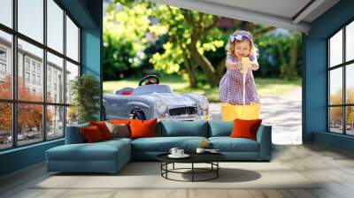 Cute gorgeous toddler girl washing big old toy car in summer garden, outdoors. Happy healthy little child cleaning car with soap and water, having fun with splashing and playing with sponge. Wall mural