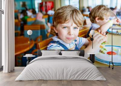 Cute healthy preschool boy eats hamburger sitting in school canteen Wall mural