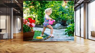 Cute little baby girl in colorful swimsuit watering plants and blossoming flowers in domestic garden on hot summer day. Adorable toddler child having fun with playing with water and ca Wall mural