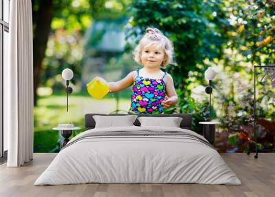 Cute little baby girl in colorful swimsuit watering plants and blossoming flowers in domestic garden on hot summer day. Adorable toddler child having fun with playing with water and can Wall mural