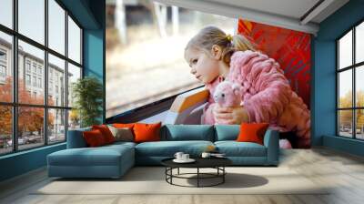 Cute little toddler girl sitting in train and looking out of window while moving. Adorable happy healthy baby child holding plush toy in hands. Smiling child going on family vacations by railroad Wall mural