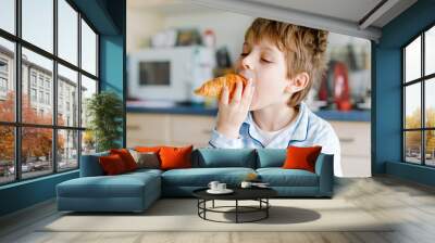 Happy little blond kid boy eating fresh croissant for breakfast or lunch. Healthy eating for children. Child in colorful pajama sitting in domestic kitchen after sleeping in the morning. Wall mural