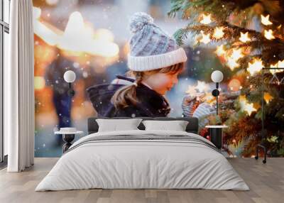 Little cute kid girl having fun on traditional Christmas market during strong snowfall. Happy child enjoying traditional family market in Germany. Schoolgirl standing by illuminated xmas tree. Wall mural