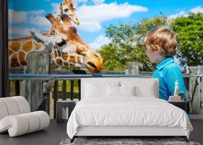 Little kid boy watching and feeding giraffe in zoo Wall mural