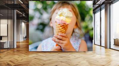 Little preschool girl eating ice cream in waffle cone on sunny summer day. Happy toddler child eat icecream dessert. Sweet food on hot warm summertime days. Bright light, colorful ice-cream Wall mural