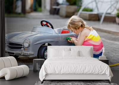 Little toddler girl playing with big vintage toy car and having fun outdoors in summer. Cute child refuel car with water. Girl using garden hose and fill up with gasoline, role game gas station. Wall mural