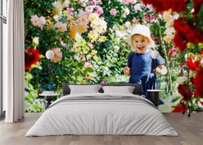 Portrait of little toddler girl in blossoming rose garden. Cute beautiful lovely child having fun with roses and flowers in a park on summer sunny day. Happy smiling baby. Wall mural