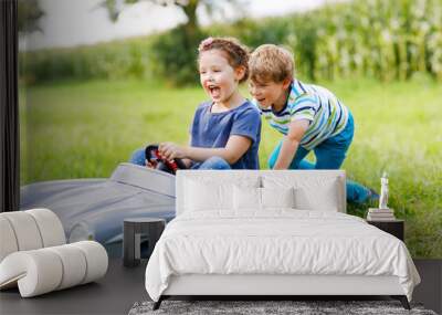 Two happy children playing with big old toy car in summer garden, outdoors. Boy driving car with little girl inside. Laughing and smiling kids. Family, childhood, lifestyle concept Wall mural