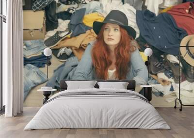 Young redhead woman in black hat and denim jacket sitting amidst a chaotic pile of clothes, looking overwhelmed. Wall mural