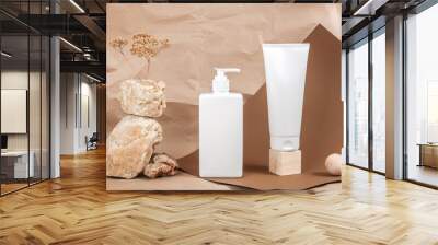 Two white tube, bottle with cream, moisturizing lotion, shampoo or other cosmetic product and stones, wooden geometric shapes, dried flowers on beige paper background. Beauty concept Wall mural