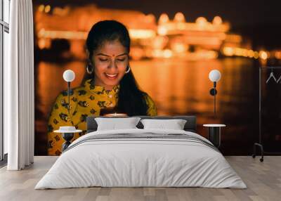 Young beautiful Indian woman holding a lit candle for Indian Festival Diwali at Ambrai Ghat in front of Picola Lake and City Palace, Udaipur, Rajasthan, India.  Wall mural