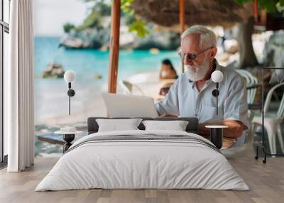 Senior man working on laptop with sea view at beach cafe. Elderly gentleman embracing technology while on tropical vacation. Retired professional using computer at seaside restaurant Wall mural