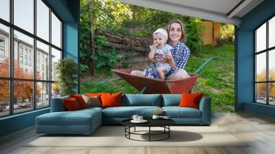 Mother and her little daughter sit in the wheelbarrow outdoors Wall mural