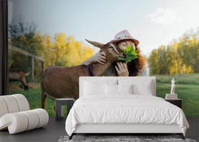 Smiling middle-aged woman in a hat bonds with the goat in peaceful rural setting. Joyful woman offers the leaves to the goat, enjoying the connection with nature on sunny day Wall mural