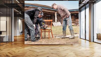 Elderly man in a wheelchair is repairing an old electric saw in his yard. His two friends are watching attentively. Wall mural