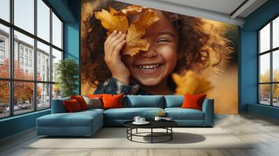 A joyful child playfully covering her eye with golden autumn leaves in a warm and vibrant outdoor setting during fall Wall mural