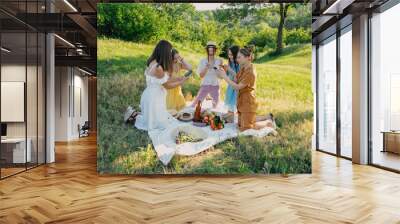 Friends having picnic in the countryside. Group of young women sitting on blanket in park near trees, at sunset on spring summer day. Five girlfriends eating and drinking red wine on outdoor party Wall mural