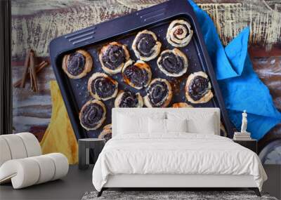 Top view: a baking tray with baked rolls with poppy seeds on a cutting board, three sticks of cinnamon, a sugar bowl with lump sugar and two kitchen napkins (yellow and blue) on a multi-colored table Wall mural