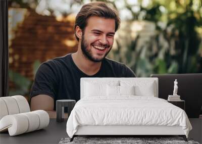 A man is sitting at a table with a laptop in front of him, smiling as he works Wall mural