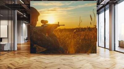 A man is sitting in a field with a gun and a camera. The sun is setting in the background Wall mural