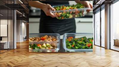 A woman is holding a tray of food in a kitchen. The tray contains a variety of vegetables, including broccoli, carrots, and peppers. The woman is wearing a black apron and she is preparing a meal Wall mural