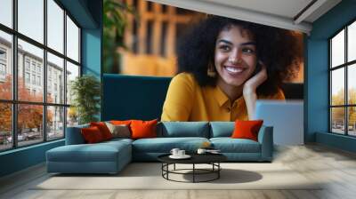 A woman with curly hair is sitting on a couch with a laptop in front of her. She is smiling and looking at the laptop screen Wall mural