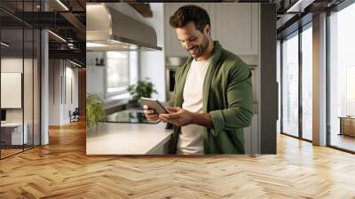 A young and handsome man uses technology at home in the kitchen with a tablet in his hands. Wall mural