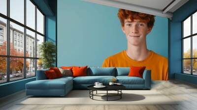 A young man with red hair is wearing a yellow shirt and smiling. He is standing in front of a blue wall Wall mural