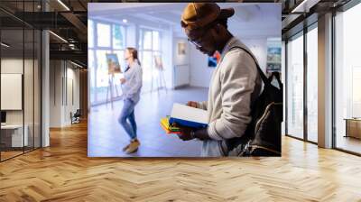 Afro-american hipster student man in eyeglasses with backpack holding colorful stack of books, reading one of book. Guy in art gallery. Multi ethnic, study abroad, culture concept. Wall mural