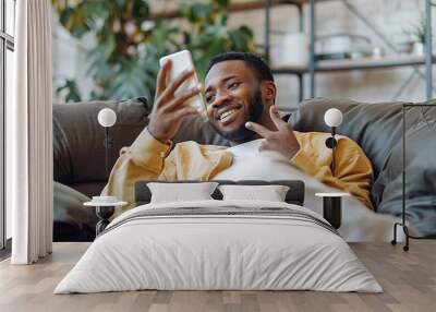 An African man relaxes on a sofa, smiling while using his phone for online communication. Wall mural