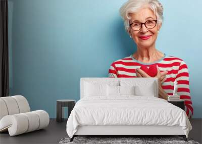 An elegant, fit granny with a charming smile, wearing glasses, holds a red apple in the kitchen. Wall mural