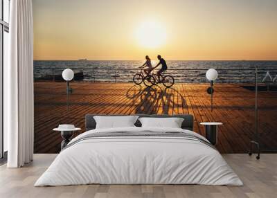 Couple of young hipsters cycling together at the beach at sunrise sky at wooden deck summer time Wall mural