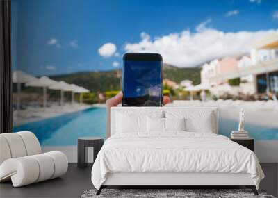 Young woman makes photo of swimming pool area, blue sky and beautiful landscape at background on smartphone camera, to share on internet social media through photo application for mobile device Wall mural