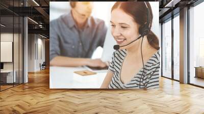 Casual dressed young woman using headset and computer while talking with customers online. Group of operators at work in sunny office. Call center Wall mural