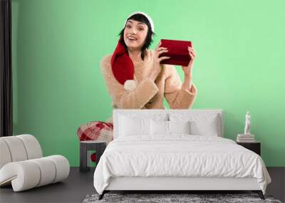 Cheerful woman in festive attire holds a red gift box against a bright green holiday background Wall mural