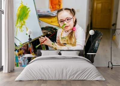 Color painting for kids with disability, child who are down syndrome, development of children with special needs. Funny little girl with down syndrome with two braids painting at easel at art class. Wall mural