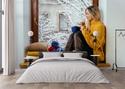 Cozy blond woman in warm yellow sweater with skeins of thread is knitting on a window sill covered with blue checkered plaid on the window at home in winter time. Wall mural