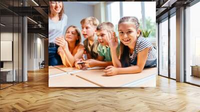 Cute enthusiastic little girl smiling, looking at camera and raising hand with teacher in class showing students a globe on background. Wall mural