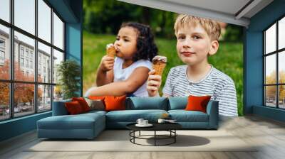 Cute little blond boy and mixed race hispanic little girl eating ice cream in green grass park, close up. Different age multiethnic children enjoying time together. Wall mural